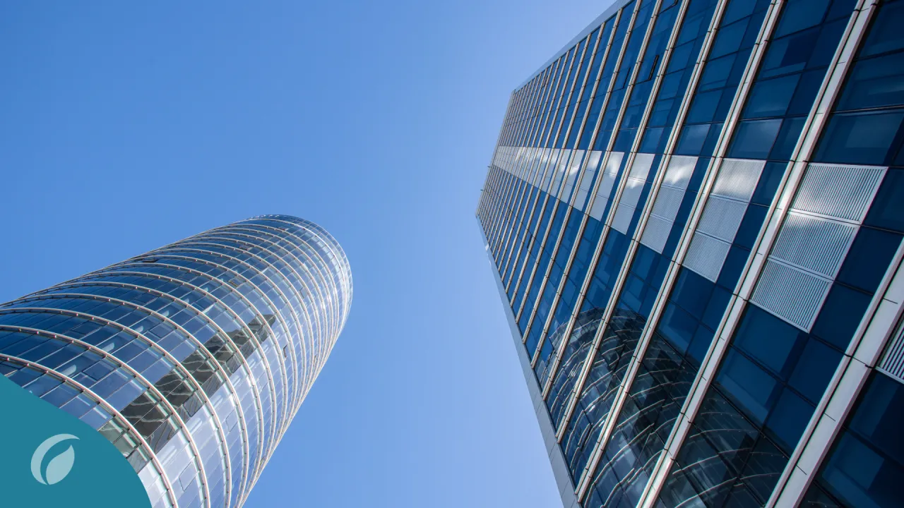 Vista desde abajo de dos edificios modernos de cristal, reflejando el cielo azul, con un logo de Investa Trust en la esquina inferior izquierda, simbolizando modernidad y protección patrimonial.