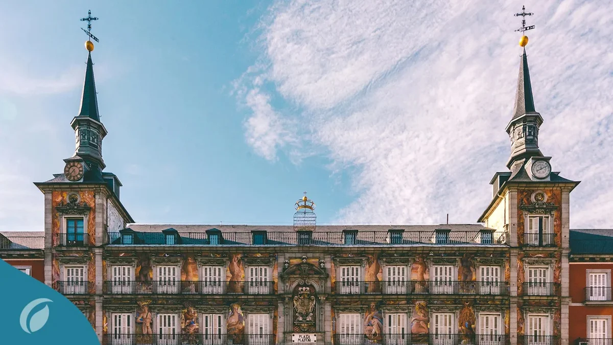 Vista de la Plaza Mayor en Madrid, destacando su arquitectura con torres y una fachada decorada. El cielo está despejado, y se aprecian detalles artísticos en la edificación.