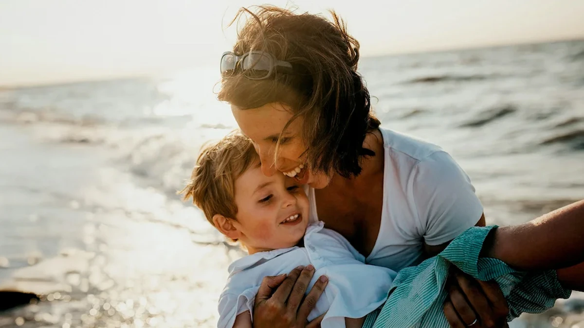 Una madre sonriente sostiene a su hijo en brazos en la playa, con el mar y la luz del atardecer de fondo, reflejando un momento de felicidad y conexión familiar.